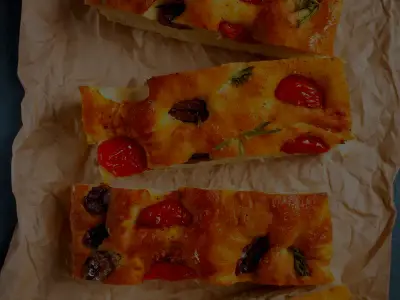 Focaccia cortada en tiras sobre papel de horno, con tomate, aceituna y romero