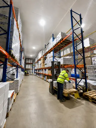 Pasillo del almacén entre estanterías de palés, con un operario preparando un pedido