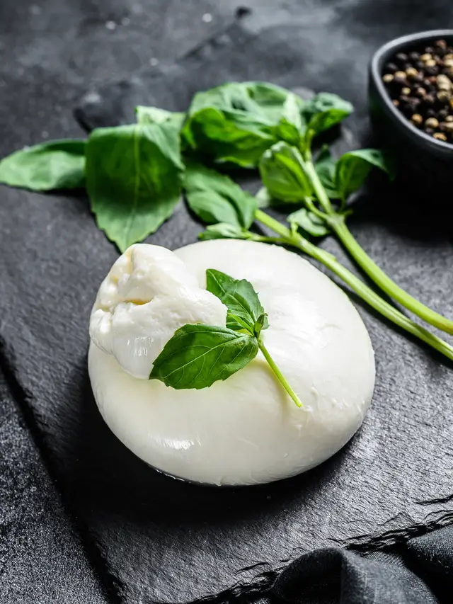 Burrata entera sobre pizarra negra, con una hoja de albahaca encima y un cuenco de pimienta en grano al lado