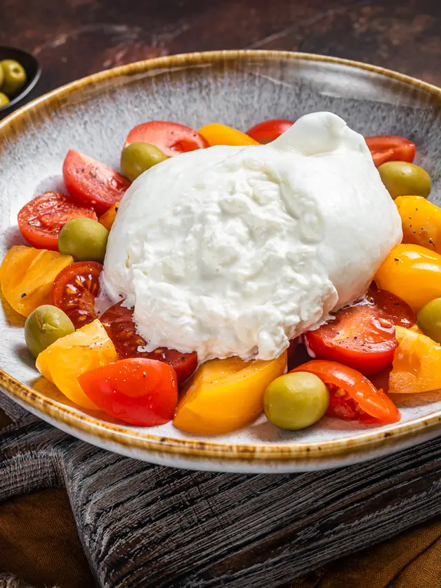 Burrata servida en plato de cerámica sobre tomate de colores y aceitunas